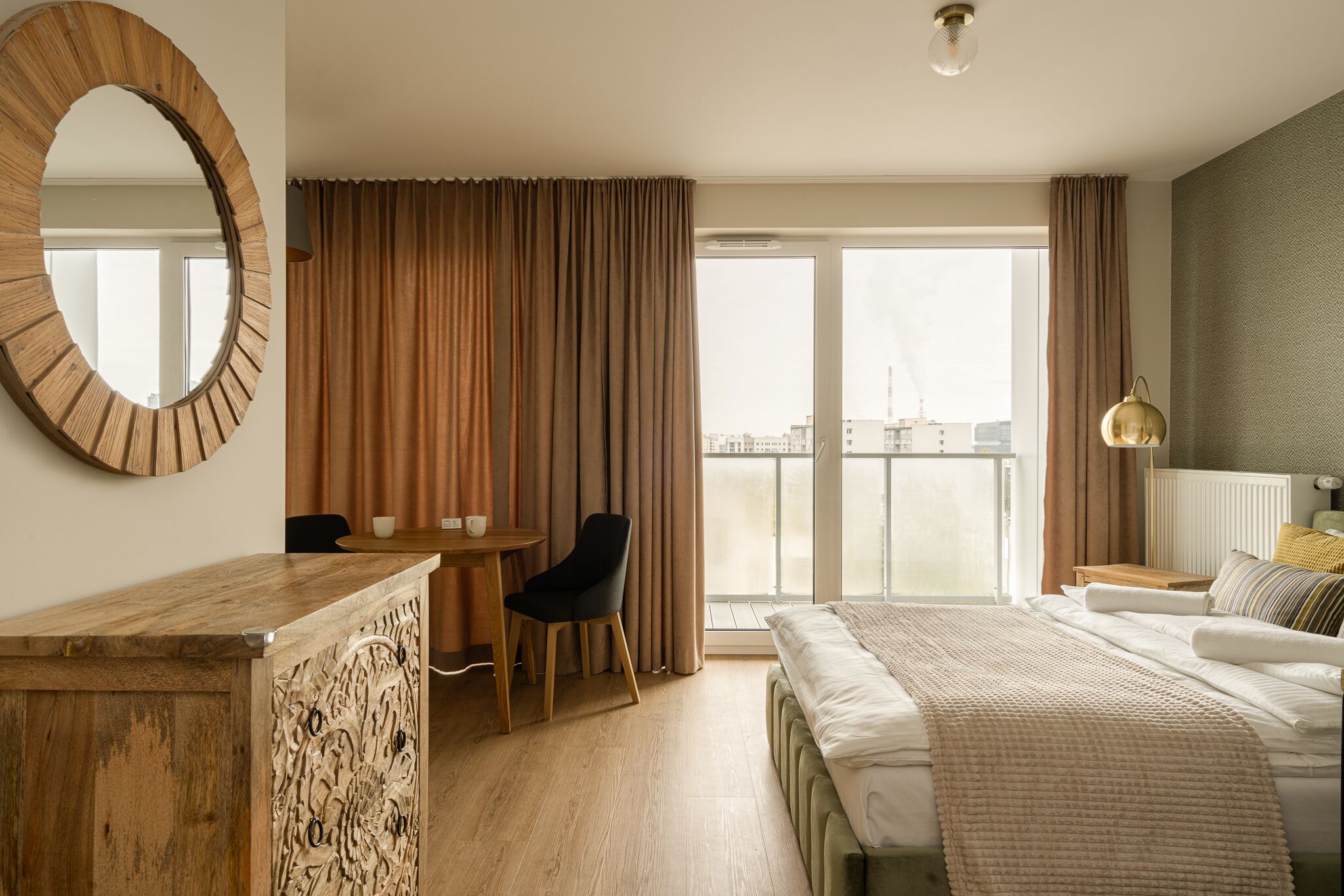 Quarto de hotel com uma cama grande, uma escrivaninha de madeira, uma cadeira, um espelho redondo e vista da cidade através de grandes janelas.