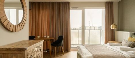 Quarto de hotel com uma cama grande, uma escrivaninha de madeira, uma cadeira, um espelho redondo e vista da cidade através de grandes janelas.