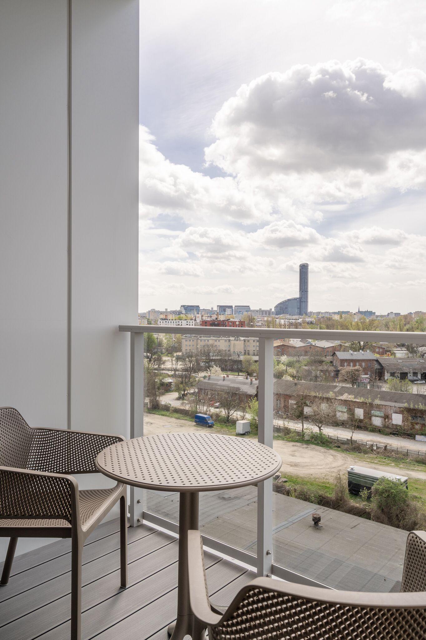 A balcony with a table and chairs, overlooking a cityscape with buildings and a clear sky.