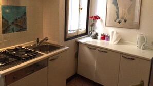 Cocina con estufa a gas, lavamanos y armarios blancos. Se observa una ventana con vista a un edificio, un cuadro en la pared y un florero con flores sobre la encimera.