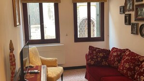 Una sala de estar con un sofá rojo, un sillón amarillo y una mesa de madera con una lámpara. Hay dos ventanas con persianas, y una pared con cuadros enmarcados.