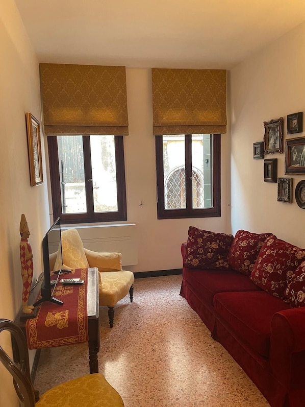 Sala de estar com um sofá vermelho, uma poltrona amarela e uma mesa de madeira com uma luminária. Há duas janelas com persianas e uma parede com quadros.