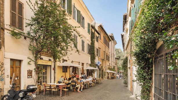 A narrow street with outdoor seating at cafes, surrounded by buildings with shutters and vines. - Luxury apartment in Trastevere (Roma)