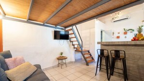 A modern living room with a sofa, a small round table, a TV mounted on the wall, a wooden staircase, and a kitchen area with a bar counter and stools. - Best Location in Sayulita - Walk to Everything (Sayulita)