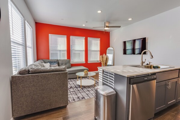 A modern kitchen with a dishwasher, a dining area with a round table, and a living room with a sofa and a TV. - Velouté Valor: 1BD Near Medical Center & Museum District (Houston)