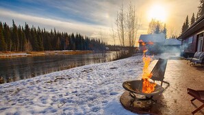 Ein mit Schnee bedeckter Deck mit Feuerstelle und Sitzgelegenheiten, mit Blick auf einen Fluss und Wald.