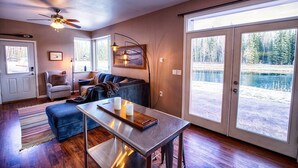 Ein gemütliches Wohnzimmer mit einer Couch, einem Couchtisch und einem Deckenventilator. Eine Tür führt zu einem Balkon mit Seeblick.