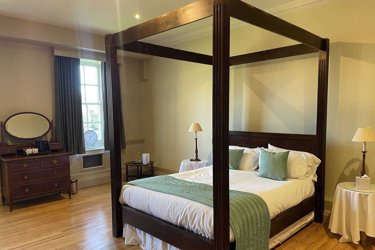 A four-poster bed with a green throw, a wooden dresser with a mirror, and a small bedside table with a lamp.