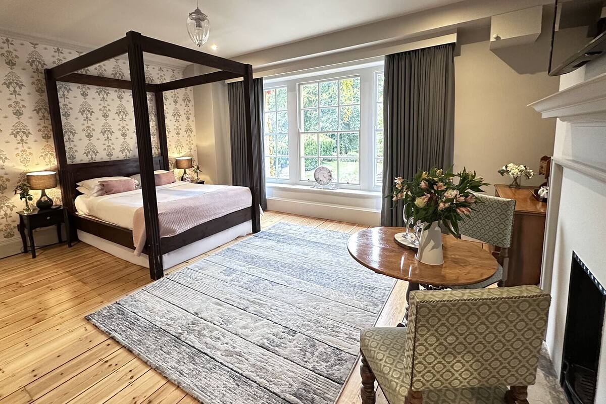 A bedroom with a four-poster bed, a wooden floor, a round table with a vase of flowers, and a view of the outdoors through a window.
