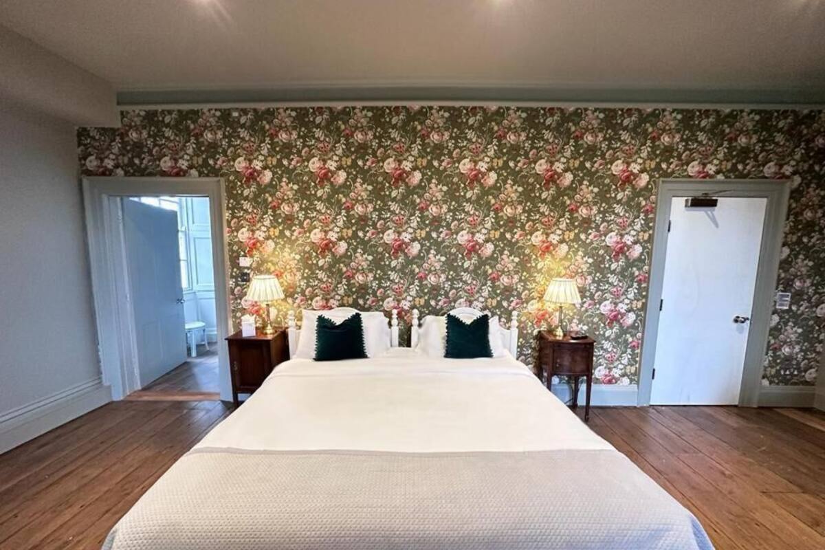 A large bed with a white bedspread and green pillows, flanked by two bedside tables with lamps. The room has wooden flooring and a floral wallpaper.