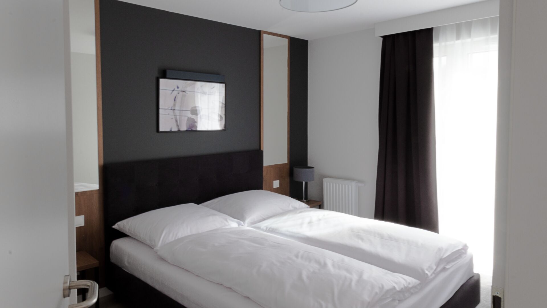 Une chambre d’hôtel avec un lit, une table de chevet, une lampe, un radiateur et une fenêtre avec des rideaux.