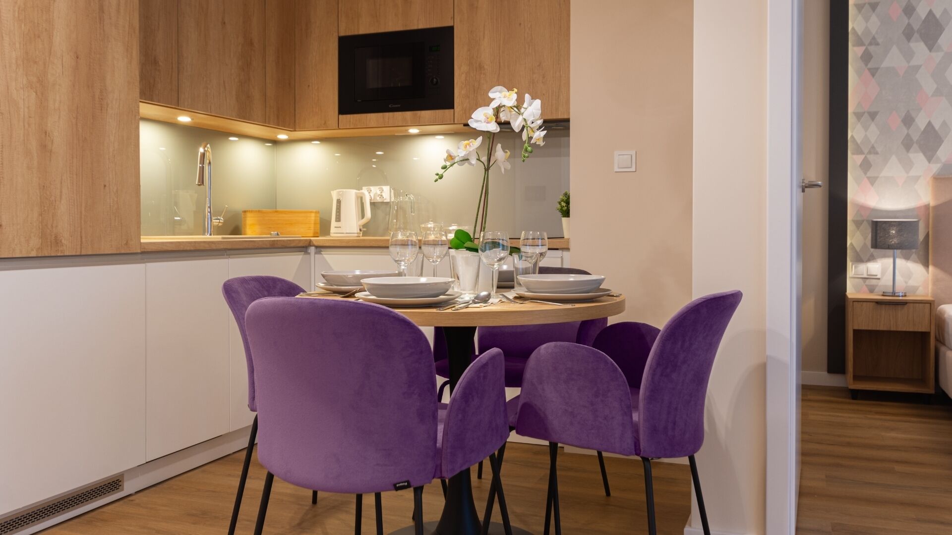 A modern kitchen with wooden cabinets, a built-in microwave, and a dining area with purple chairs and a round table set for four.