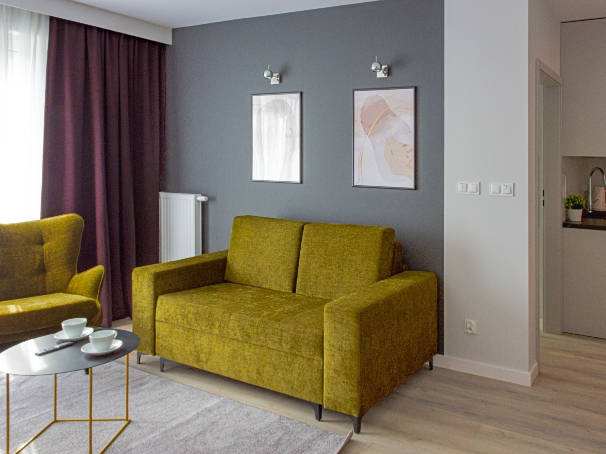 A modern living room with a green sofa, a small round table with cups, and two framed abstract paintings on the wall.