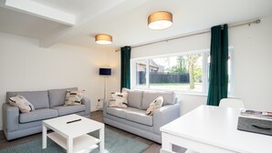 A modern living room with a grey sofa, white coffee table, and a dining table. There are two ceiling lights and a window with curtains.