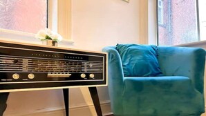 A room with a vintage radio on a white stand, a teal armchair, and a fluffy white rug. - 4 bed cottage (Contractors Welcome) Egham (Egham)