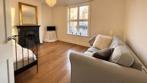 A room with a bed, a sofa, a small table, a TV, a chandelier, and a window with a view of buildings. - 4 bed cottage (Contractors Welcome) Egham (Egham)