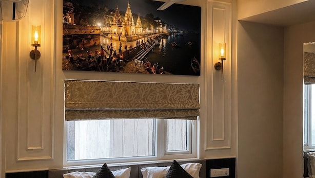 A hotel room with a bed, bedside tables, a ceiling fan, a large framed picture on the wall, and a window with blinds. - Anaya Retreat (Varanasi)
