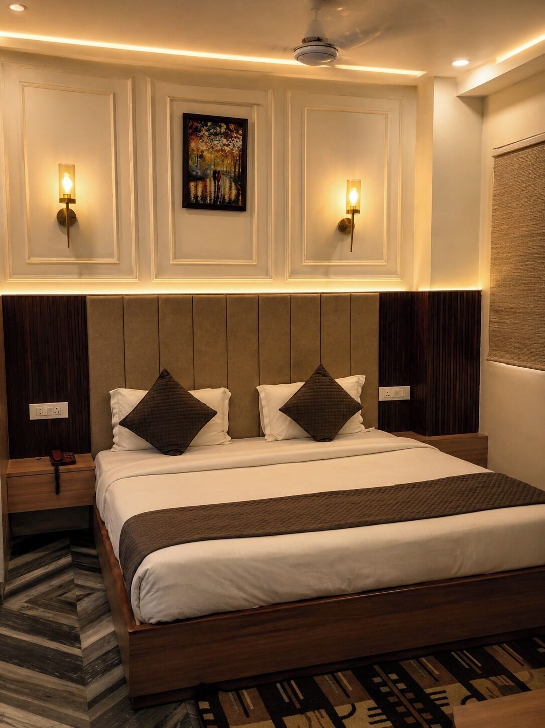 A hotel room with a large bed, two bedside tables, a ceiling fan, wall-mounted lights, and a painting above the bed.