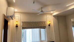 A room with a ceiling fan, recessed lighting, a window with a patterned valance, and wall-mounted sconces. - Anaya Retreat (Varanasi)