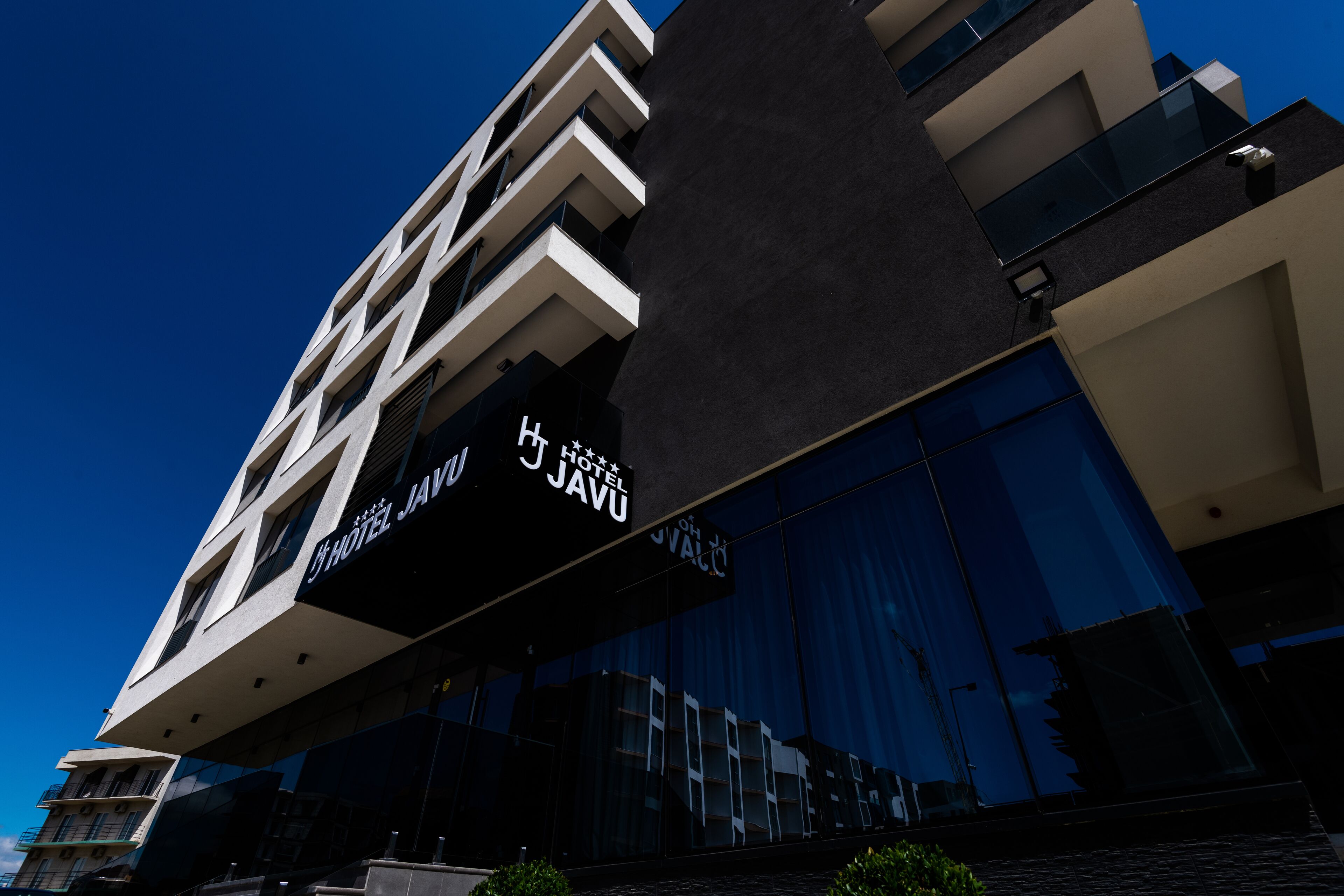 Un hôtel moderne, reconnaissable à son enseigne bien visible « Hôtel J’JAVU », une structure à plusieurs étages dotée de balcons et d’une façade en verre réfléchissant.