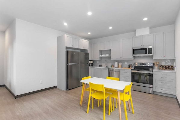 Una cocina moderna con armarios blancos, electrodomésticos de acero inoxidable y una zona de comedor con mesa blanca y sillas amarillas.