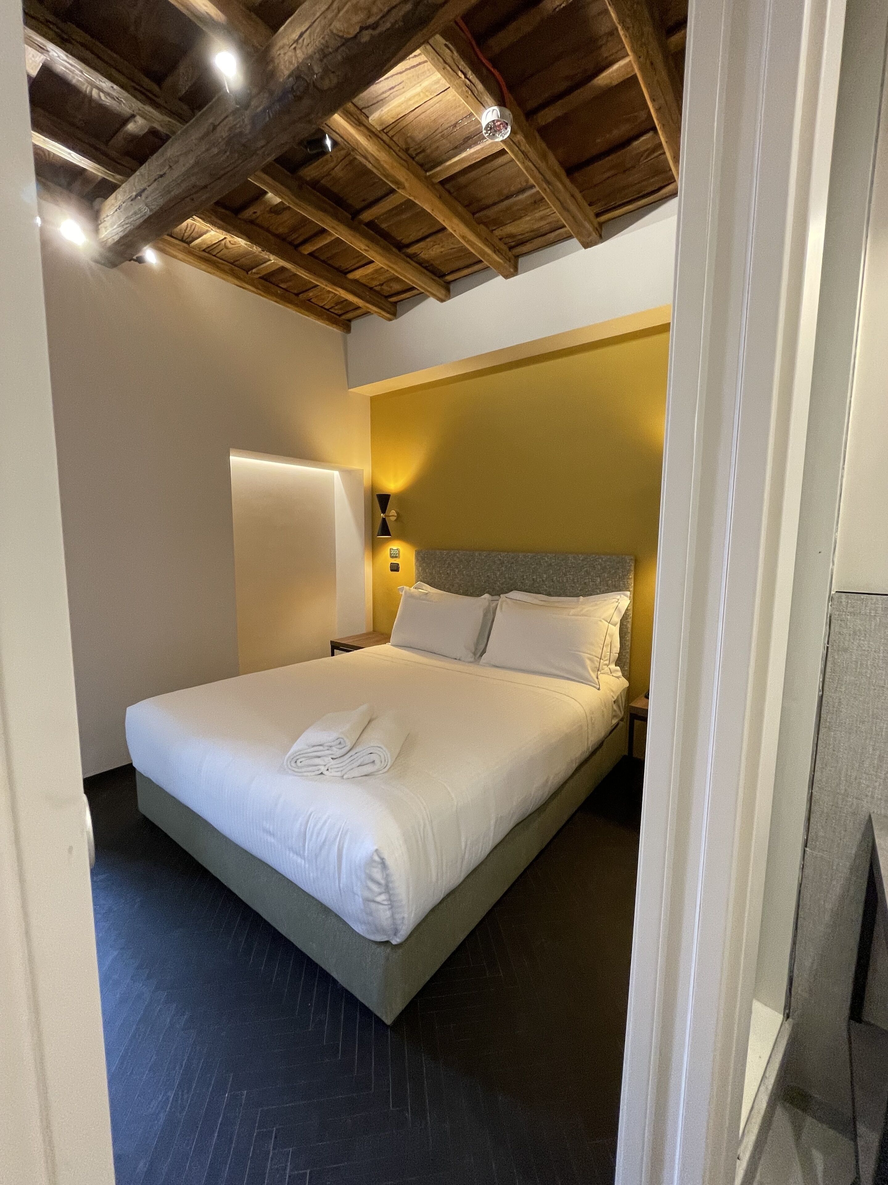A neatly made bed with white linens and a folded towel, a bedside lamp, and a wooden ceiling.