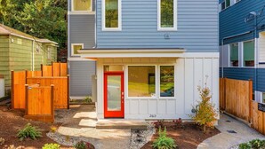 Una casa de dos pisos con puerta roja, detalles blancos y un pequeño jardín delantero con cerca de madera y plantas.