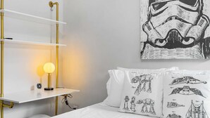 Un dormitorio con ropa de cama blanca, una almohada con temática de Star Wars, una lámpara de noche y un cartel de Star Wars en la pared.