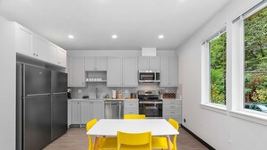 Una cocina moderna con armarios grises, electrodomésticos de acero inoxidable, una mesa de comedor blanca con sillas amarillas y una ventana con vistas a la vegetación.