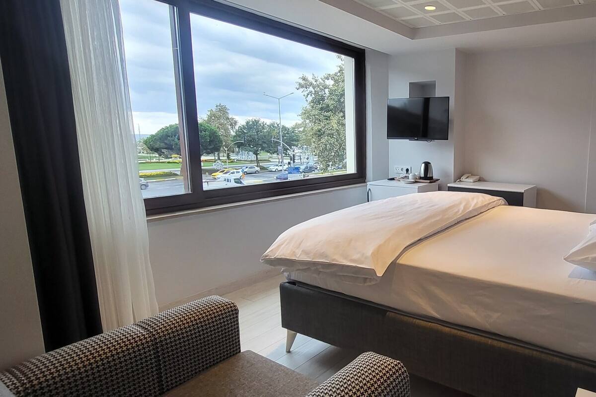 A modern hotel room with a large bed, a bedside table with a lamp, a TV mounted on the wall, and a window offering a view of a parking lot and trees.