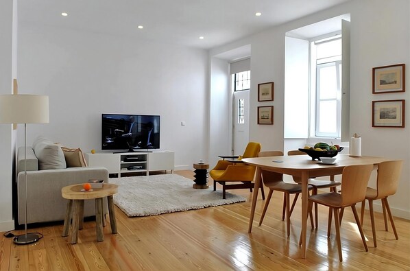 A modern living room with a flat-screen TV, a sofa, a dining table with chairs, and a small side table.