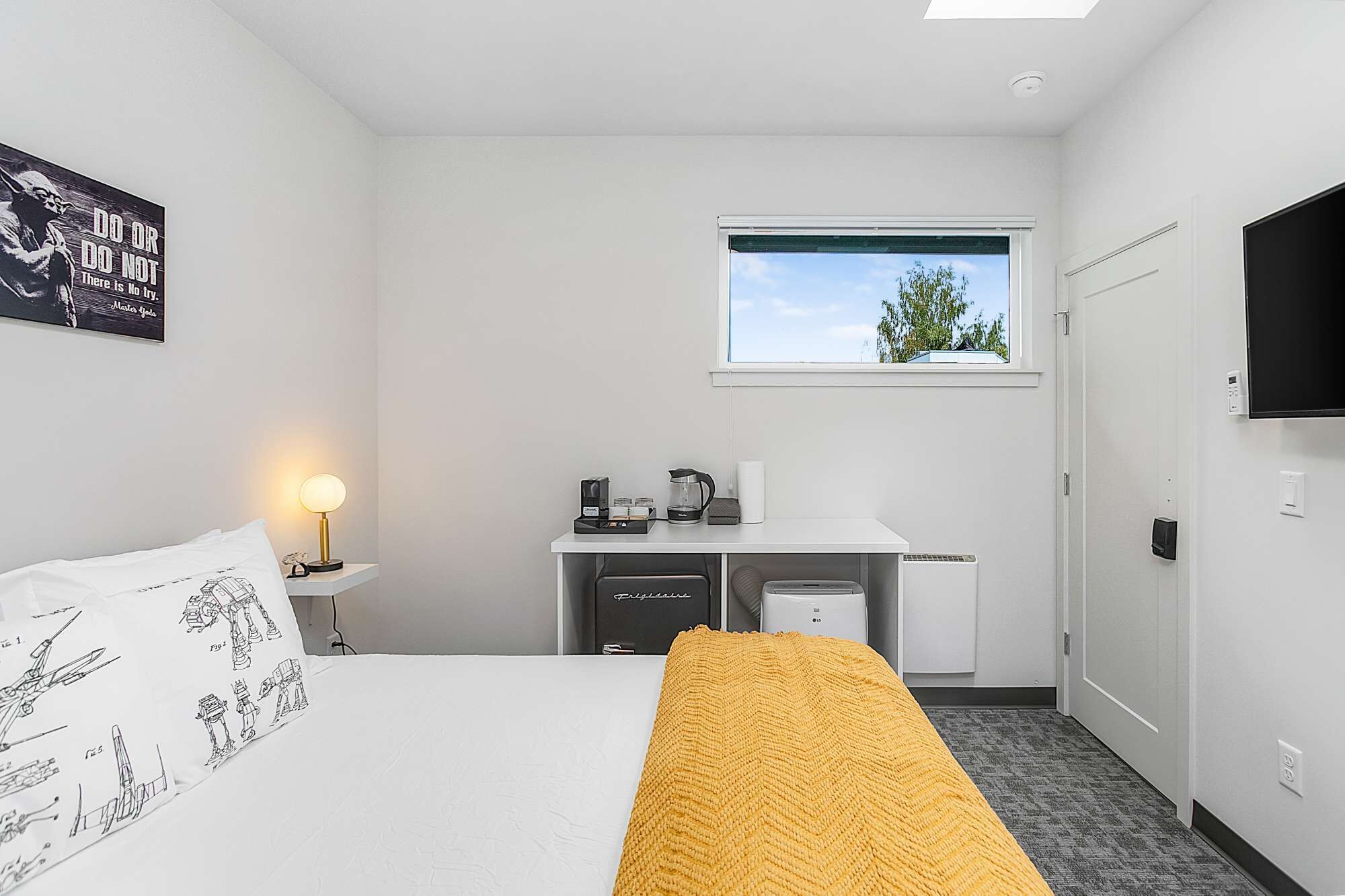 Een hotelkamer met een bed, een bureau met een computer, een raam met uitzicht op bomen en een aan de muur bevestigde televisie.