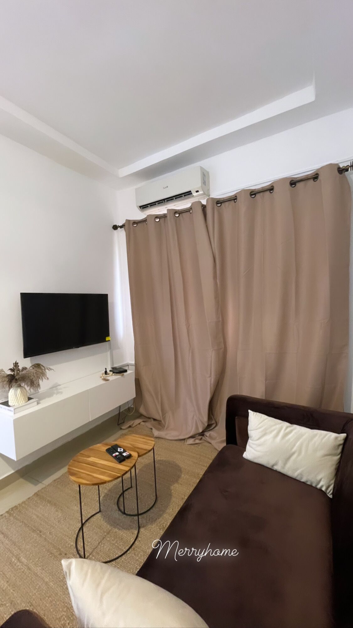 A modern living room with a flat-screen TV, a sofa, a coffee table, and a large window with beige curtains.
