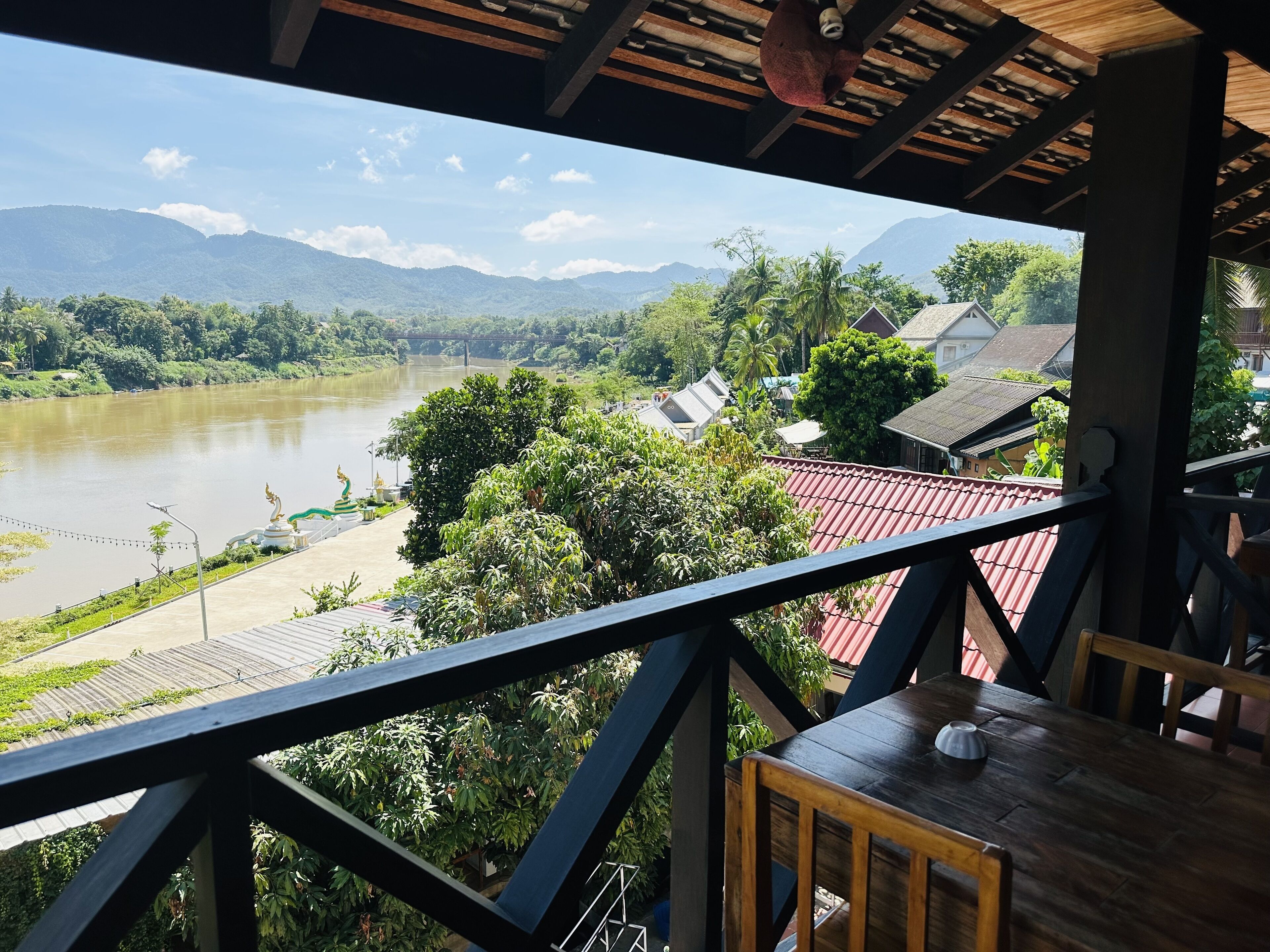 Une vue depuis un balcon donnant sur une rivière, avec des montagnes en arrière-plan, un petit pont et une statue.