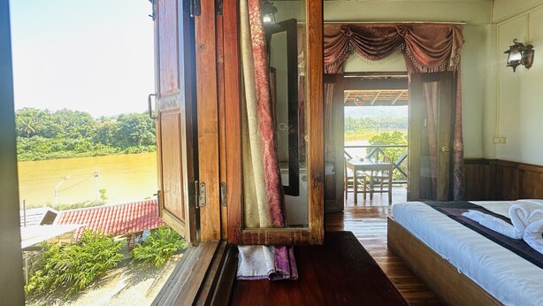 A room with a wooden floor, a bed with white bedding, a window with curtains, and a view of a green landscape. - Kingkham Riverside Hotel (Luang Prabang)