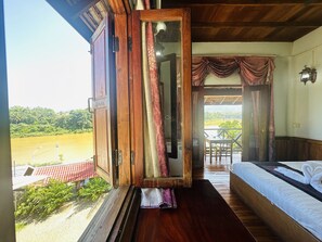 Una habitación con piso de madera, cama con ropa de cama blanca, ventana con cortinas y vista a un paisaje verde.