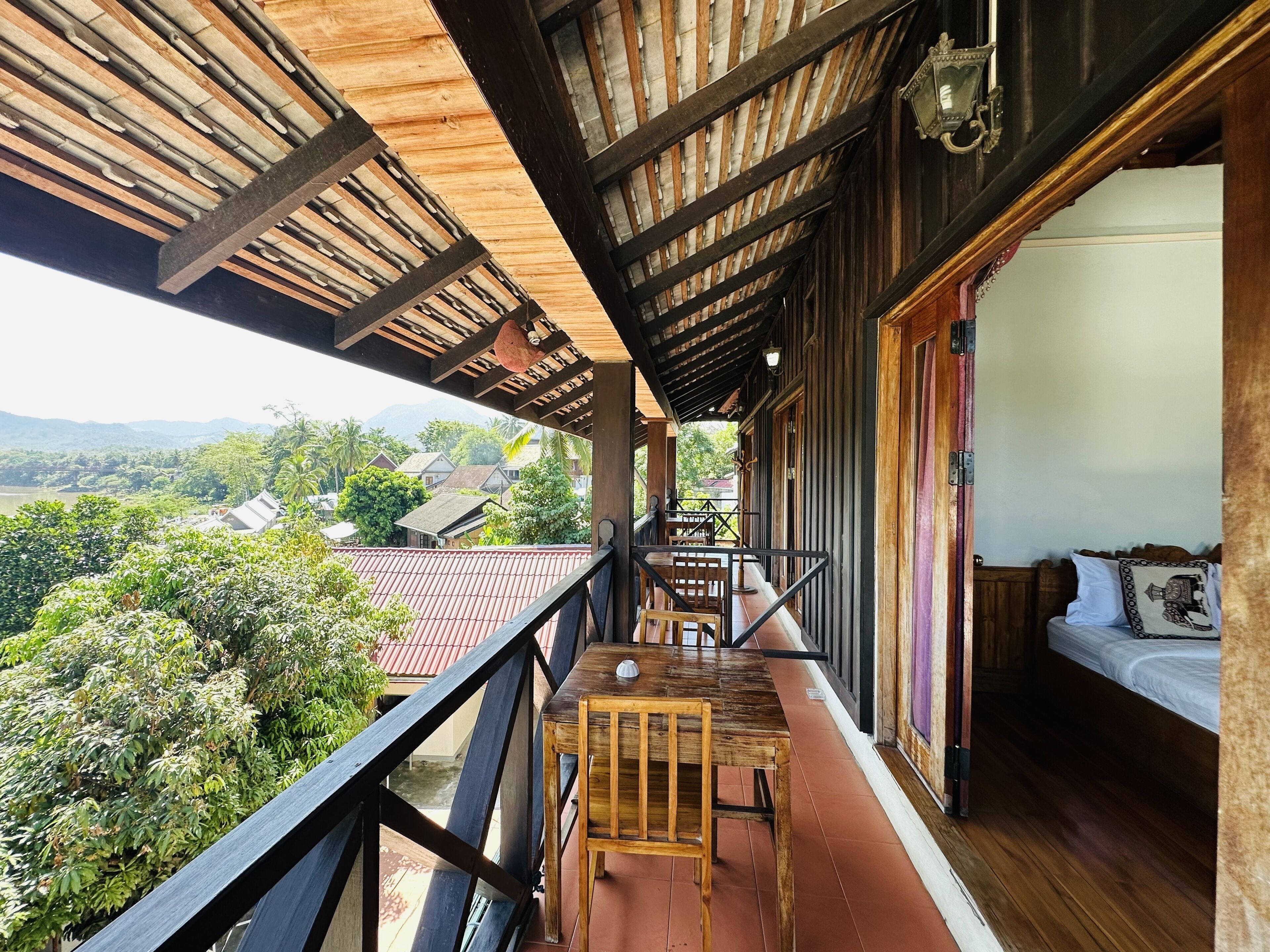 Un balcon avec une table et des chaises en bois, un lit avec un oreiller et une vue sur un village et des montagnes.