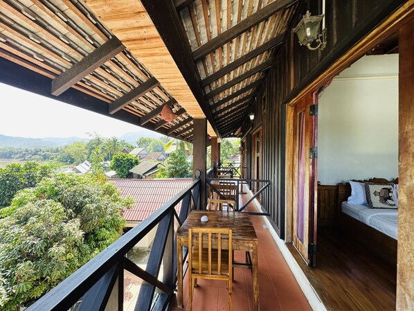 Un balcón con una mesa y sillas de madera, una cama con almohada, y vistas a un pueblo y montañas.