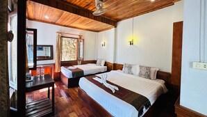 A hotel room with two beds, wooden flooring, a desk with a mirror, and a stone wall. - Kingkham Riverside Hotel (Luang Prabang)