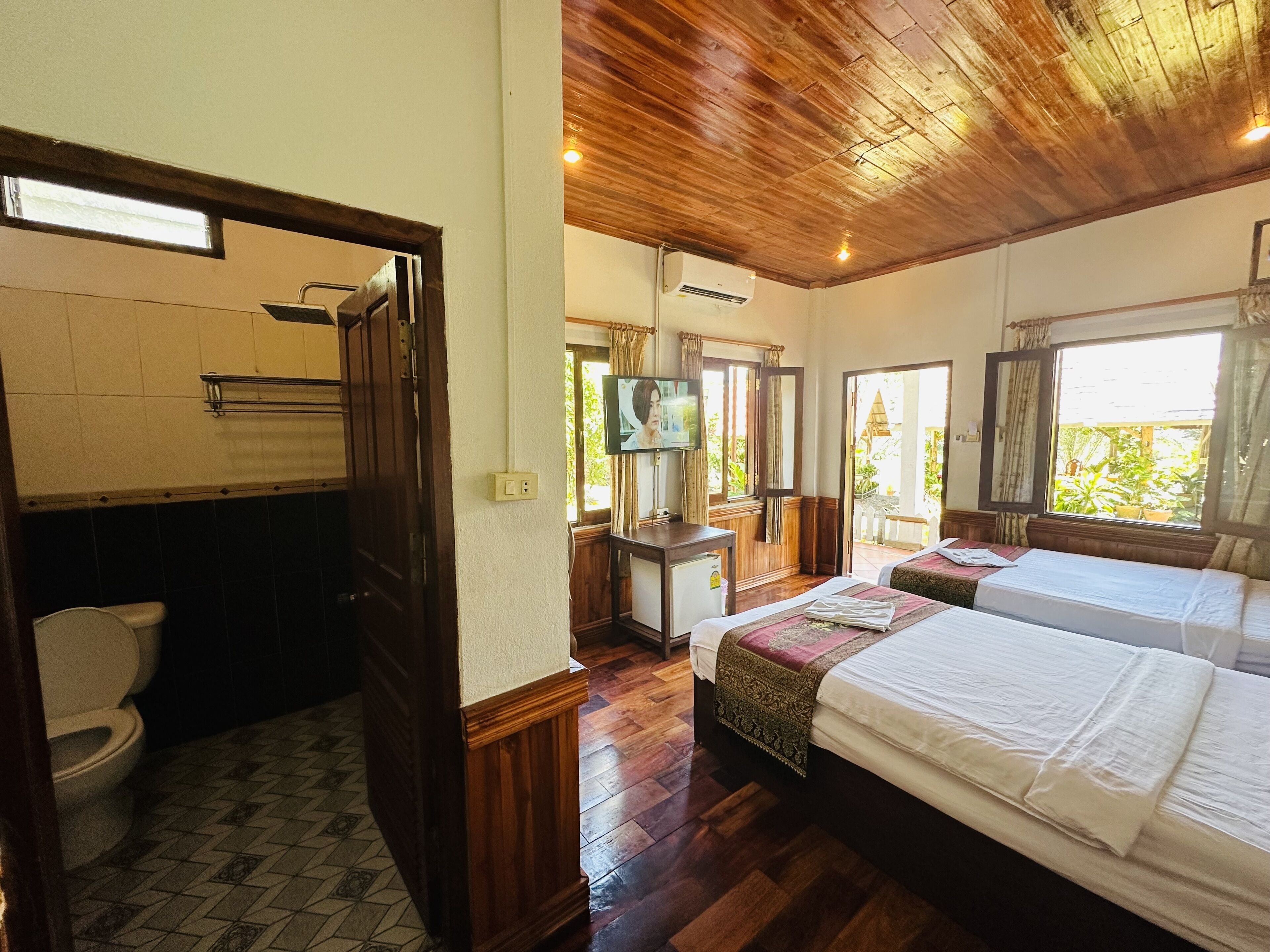 Une chambre d’hôtel avec un plafond en bois, une salle de bain avec des toilettes et un lavabo, deux lits avec du linge de lit blanc, un téléviseur posé sur un meuble en bois et une fenêtre avec des rideaux.