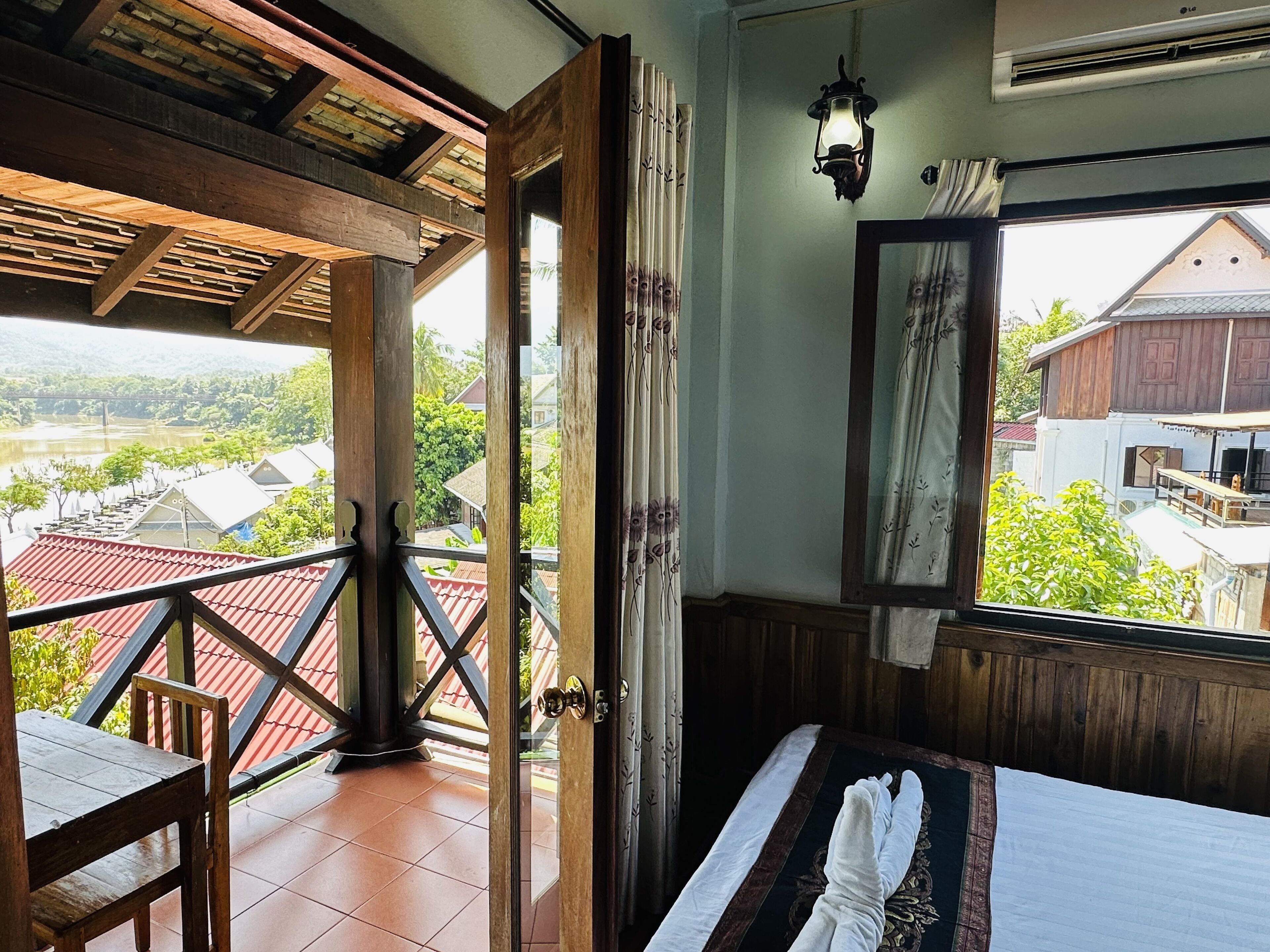 A room with a balcony, a bed with a towel, a wooden table, and a view of houses and greenery outside.