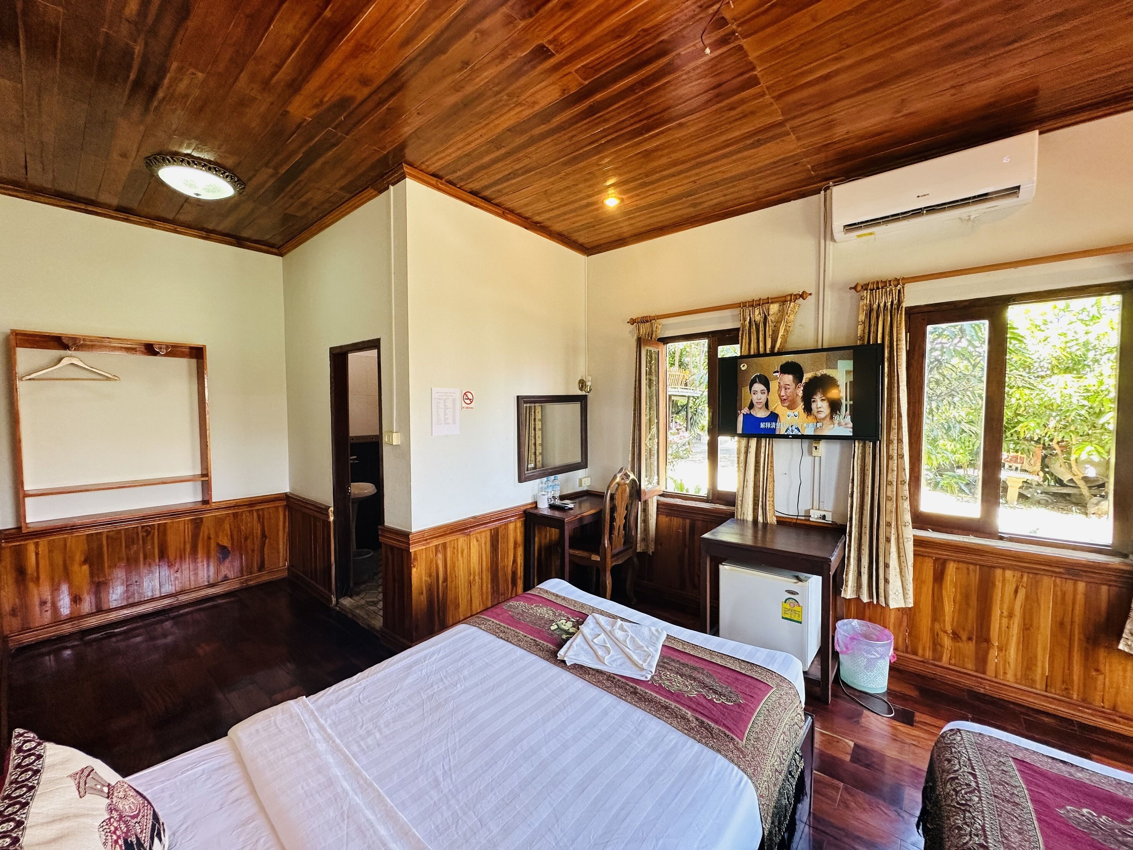 Habitación de hotel con techo de madera, cama con colcha blanca y roja, escritorio con televisor y ventana con cortinas.