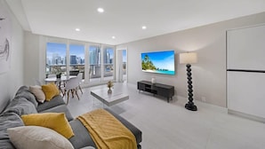 A modern living room with a sofa, coffee table, TV, and a city view through large windows. - Bayfront High-Rise Condo | Oceanview | StayCozy (Miami)