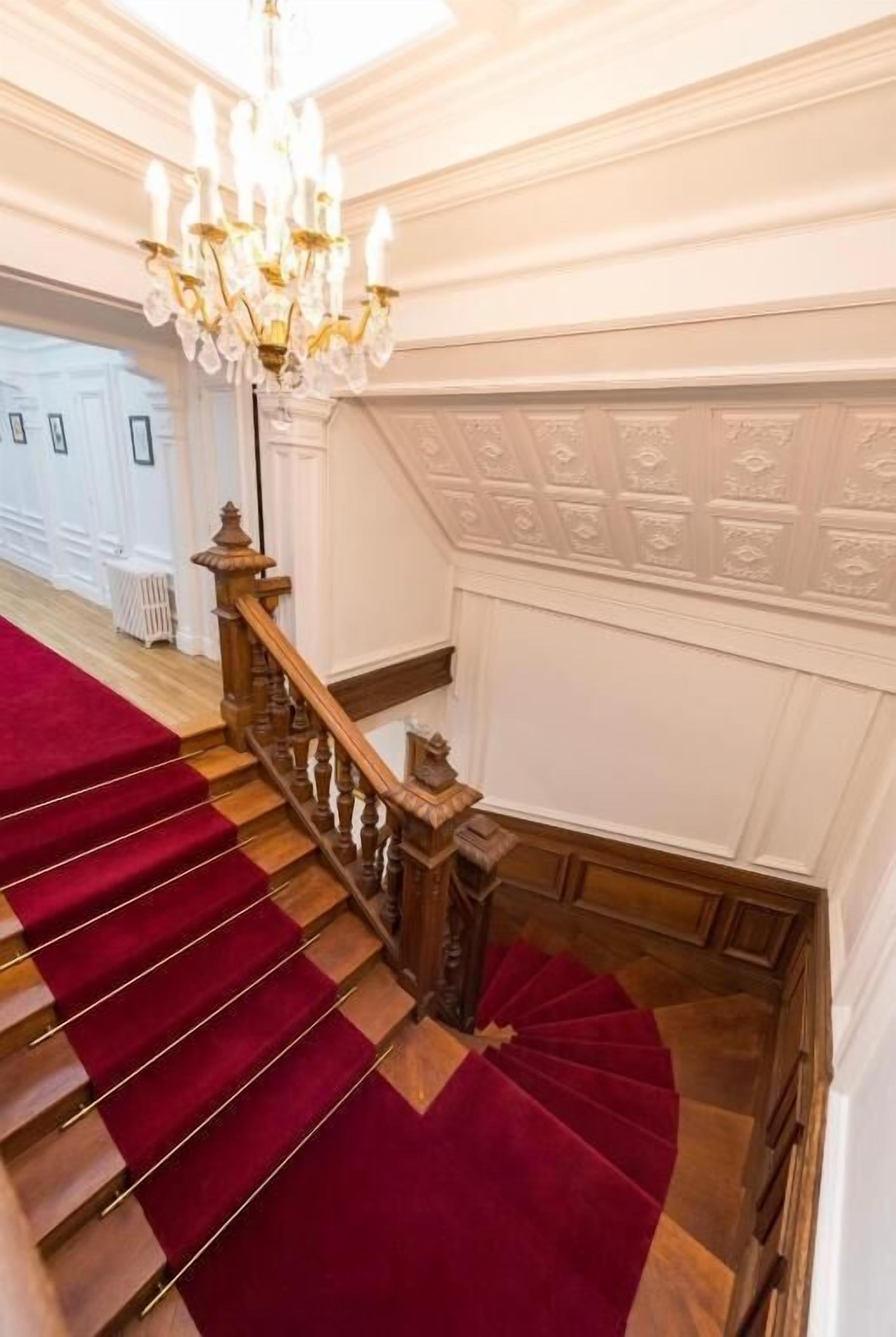 A grand staircase with a red carpet, wooden balustrade, and a chandelier.