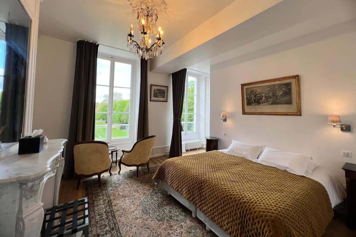 Habitación de hotel con cama, dos sillas, escritorio, candelabro y ventana con cortinas.