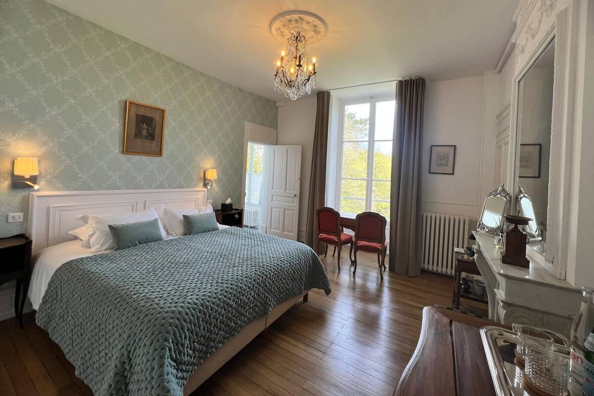 Habitación de hotel con una cama grande, una araña de cristal, una mesa de comedor con sillas y vistas al exterior a través de una ventana.