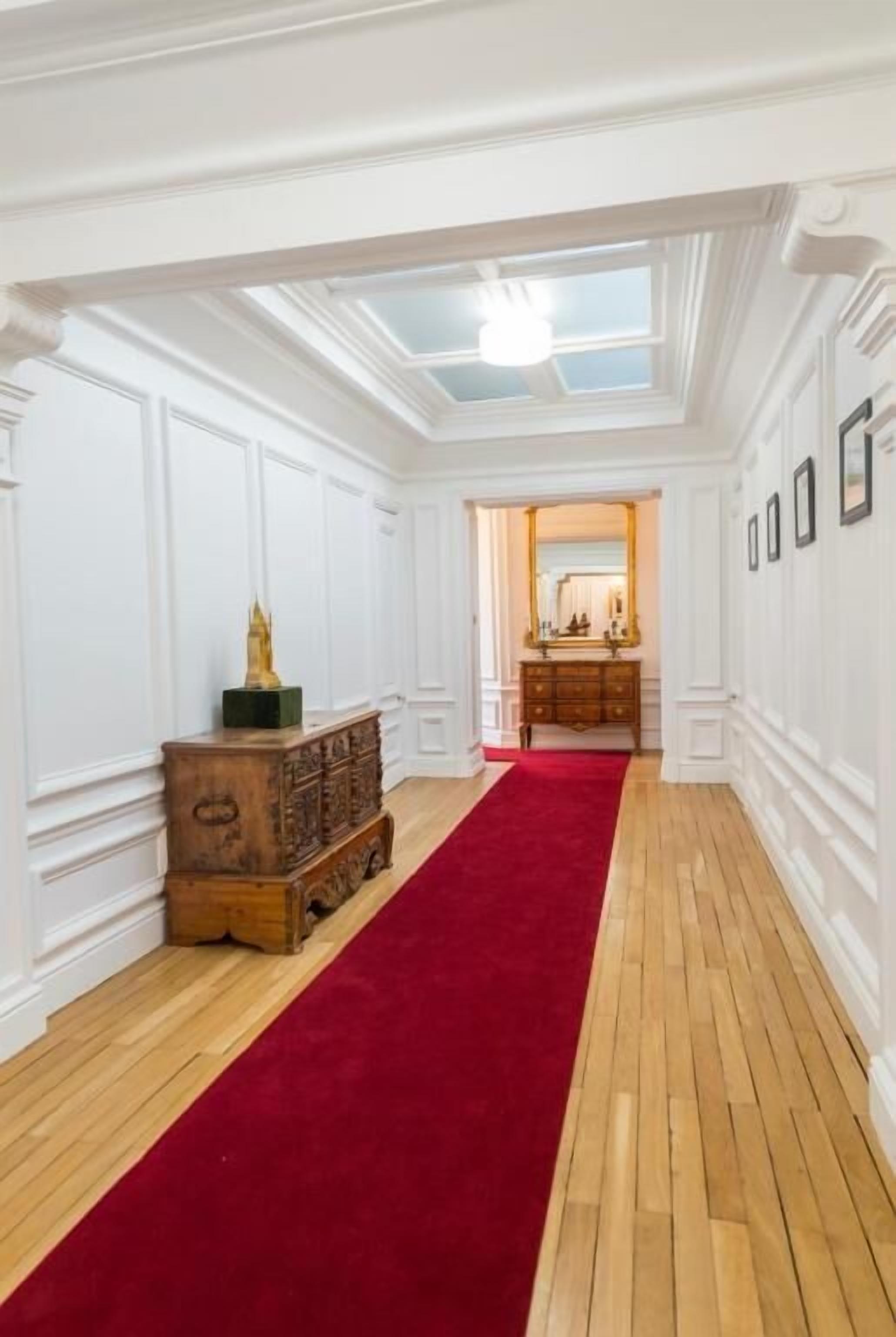 Un pasillo con alfombra roja, suelo de madera, paredes blancas y un techo con tragaluces.