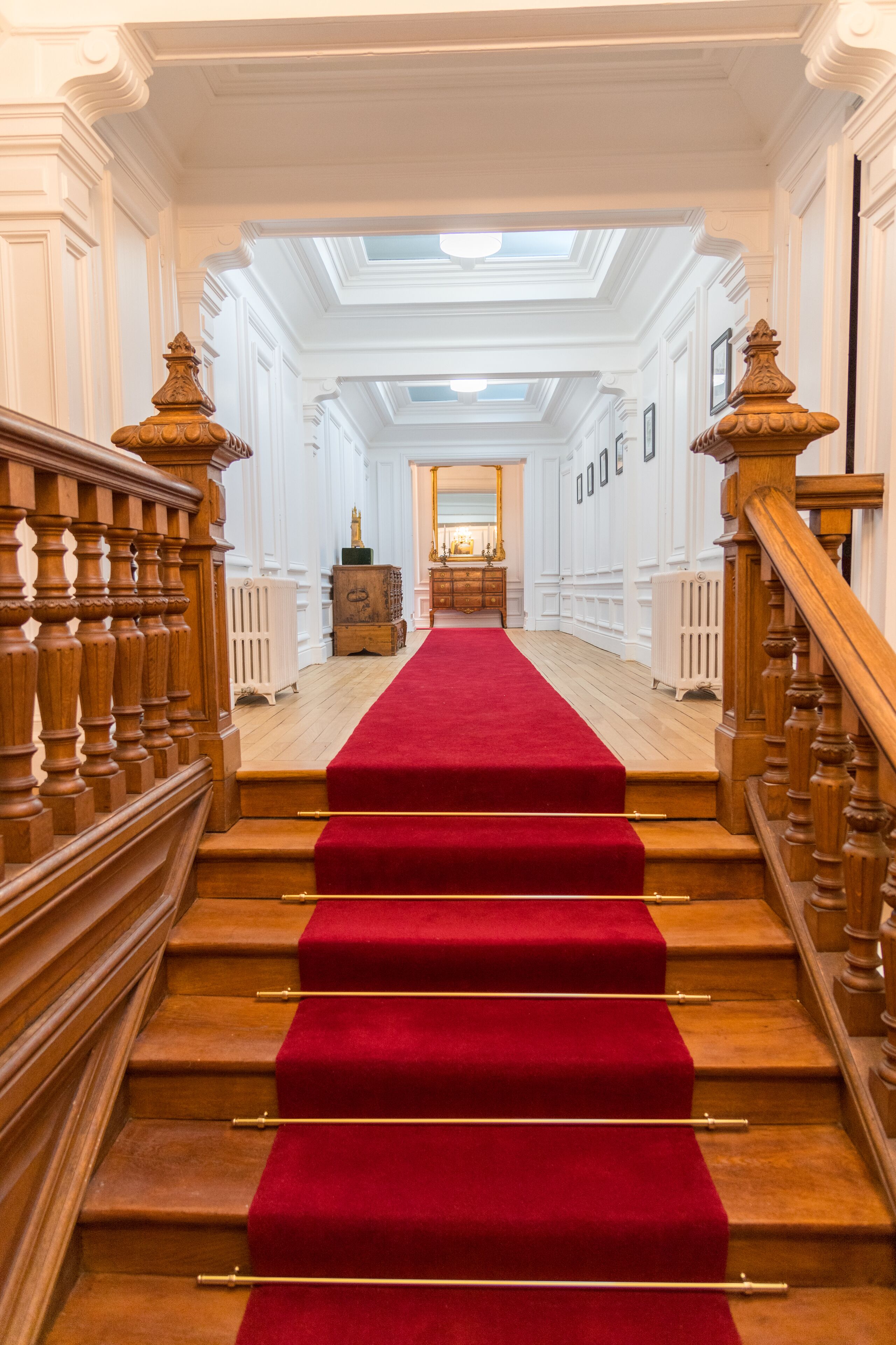 A grand staircase with a red carpet, flanked by wooden balusters and a white wall with framed pictures.
