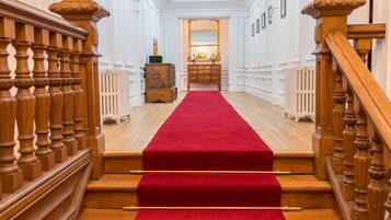A grand staircase with a red carpet, flanked by wooden balusters and a white wall with framed pictures.