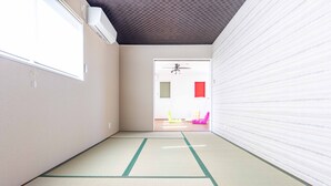 Una habitación con suelo de esteras de tatami, ventilador de techo y puerta corrediza que da a otra habitación con sillas de colores.